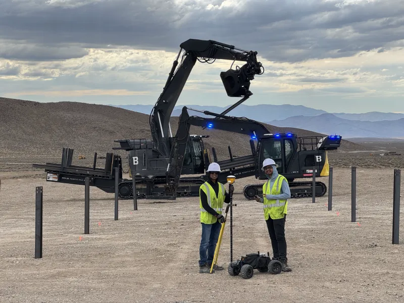 Two engineers in hard hats with a GNSS survey pole and the survey robot in front of robotic pile drivers